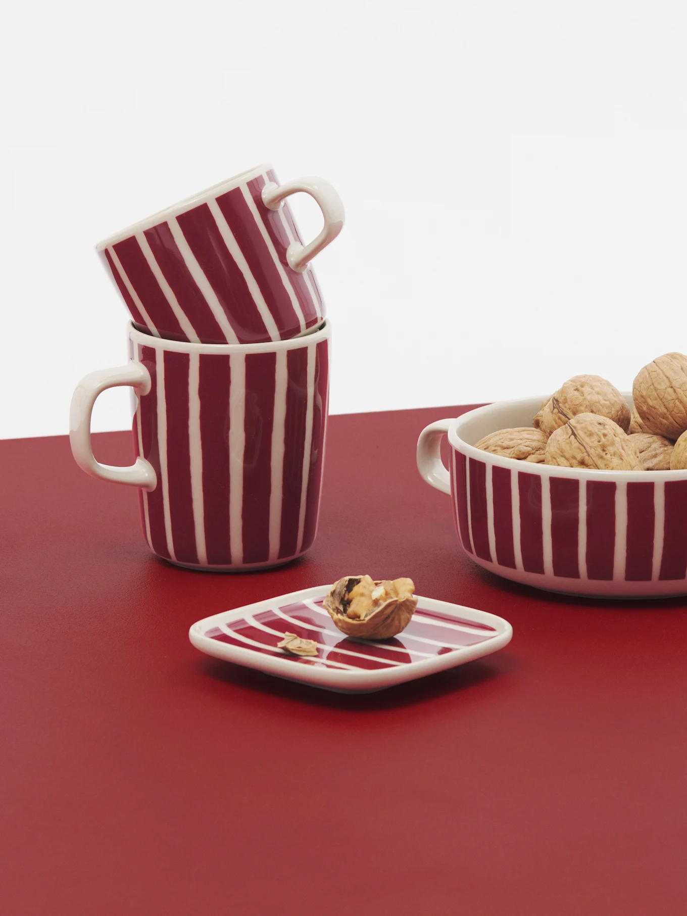 Red and white striped mugs and a bowl of walnuts on a red surface with a white background.