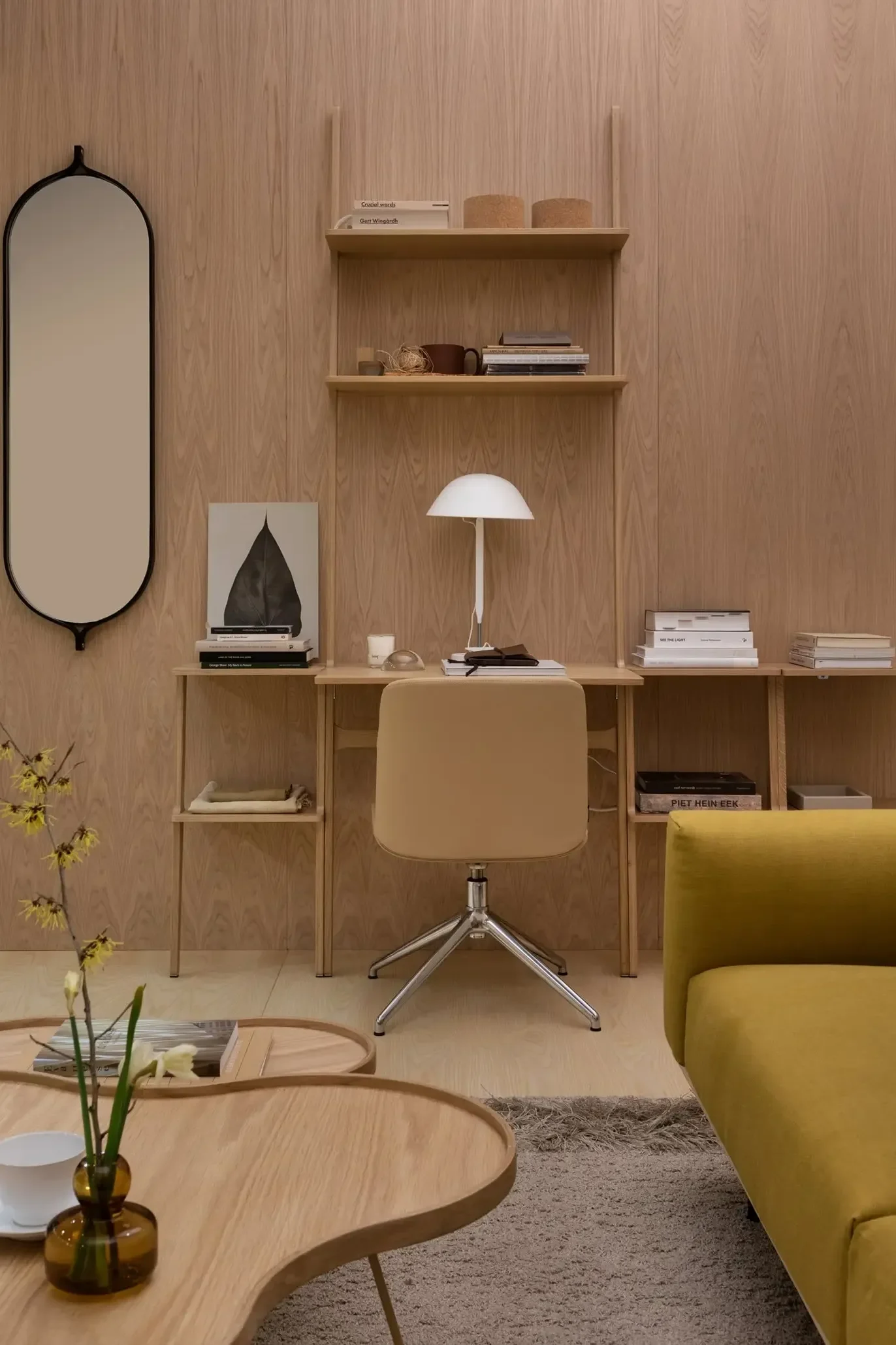 A minimalist home office with light wood paneling, a beige desk chair, white lamp, and shelves, next to a yellow sofa and coffee tables.