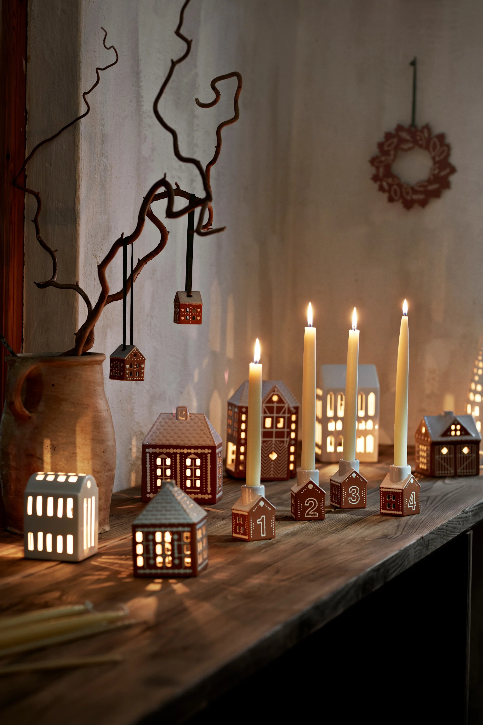 Cozy Advent display with lit candles in numbered gingerbread house holders, house lanterns, and hanging ornaments on a branch.