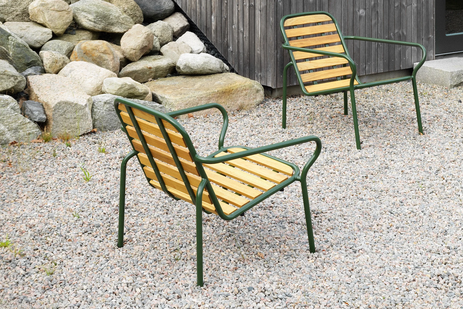 Two modern outdoor chairs with green frames and wooden slats on a gravel patio, next to a rock wall and a wooden building.