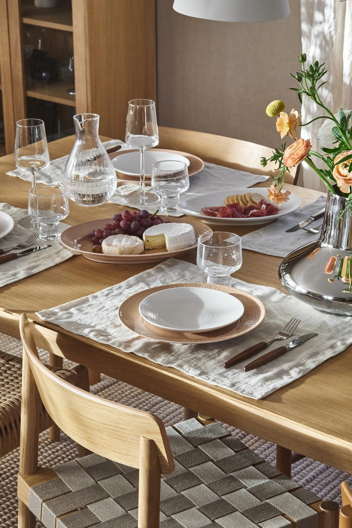 A wooden dining table set with plates, glasses, a water carafe, grapes, cheeses, cured meats, and flowers. Woven chairs.