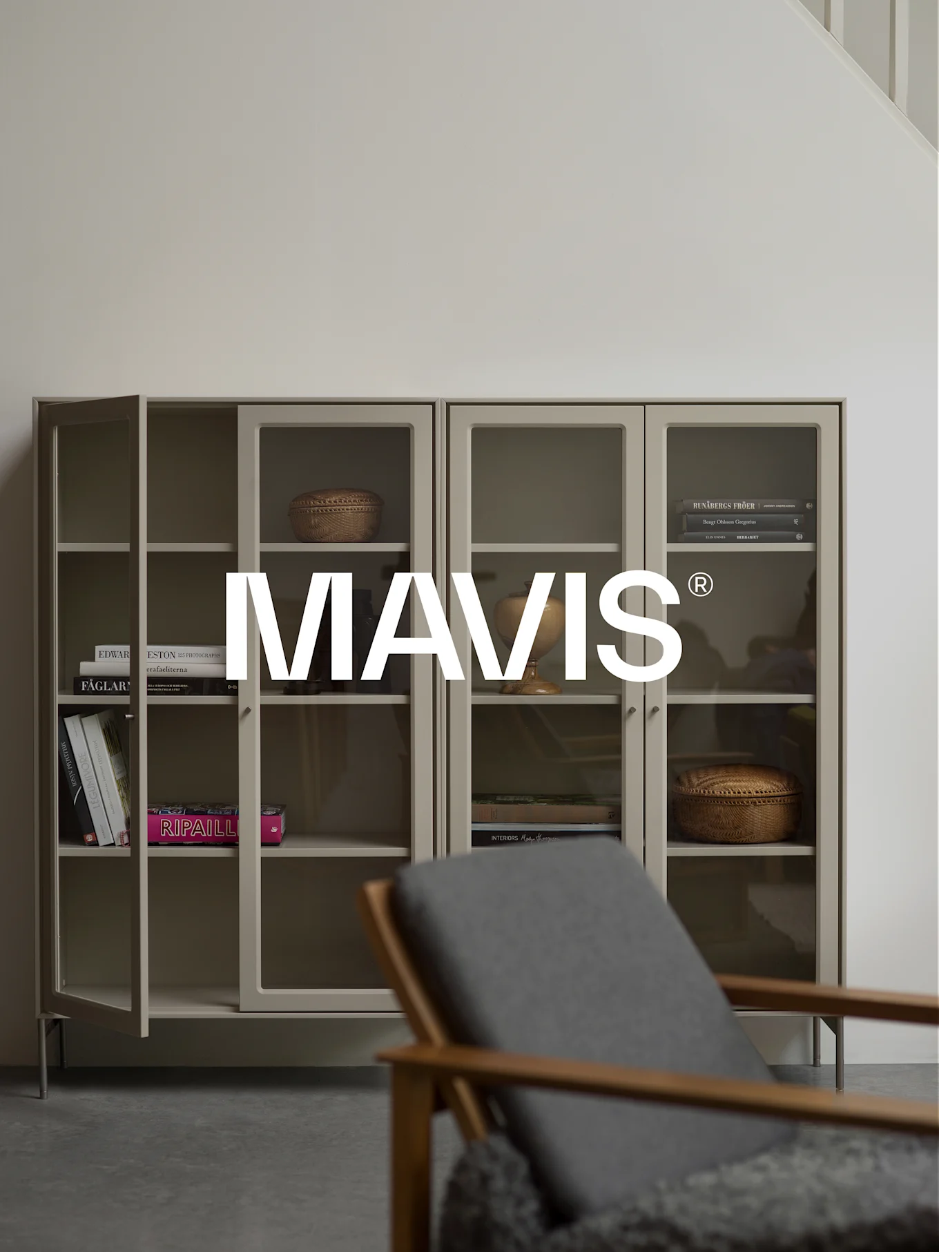 Two light grey cabinets with glass doors, displaying books and decor. A wooden chair is blurred in the foreground. 'MAVIS®' logo is centered.