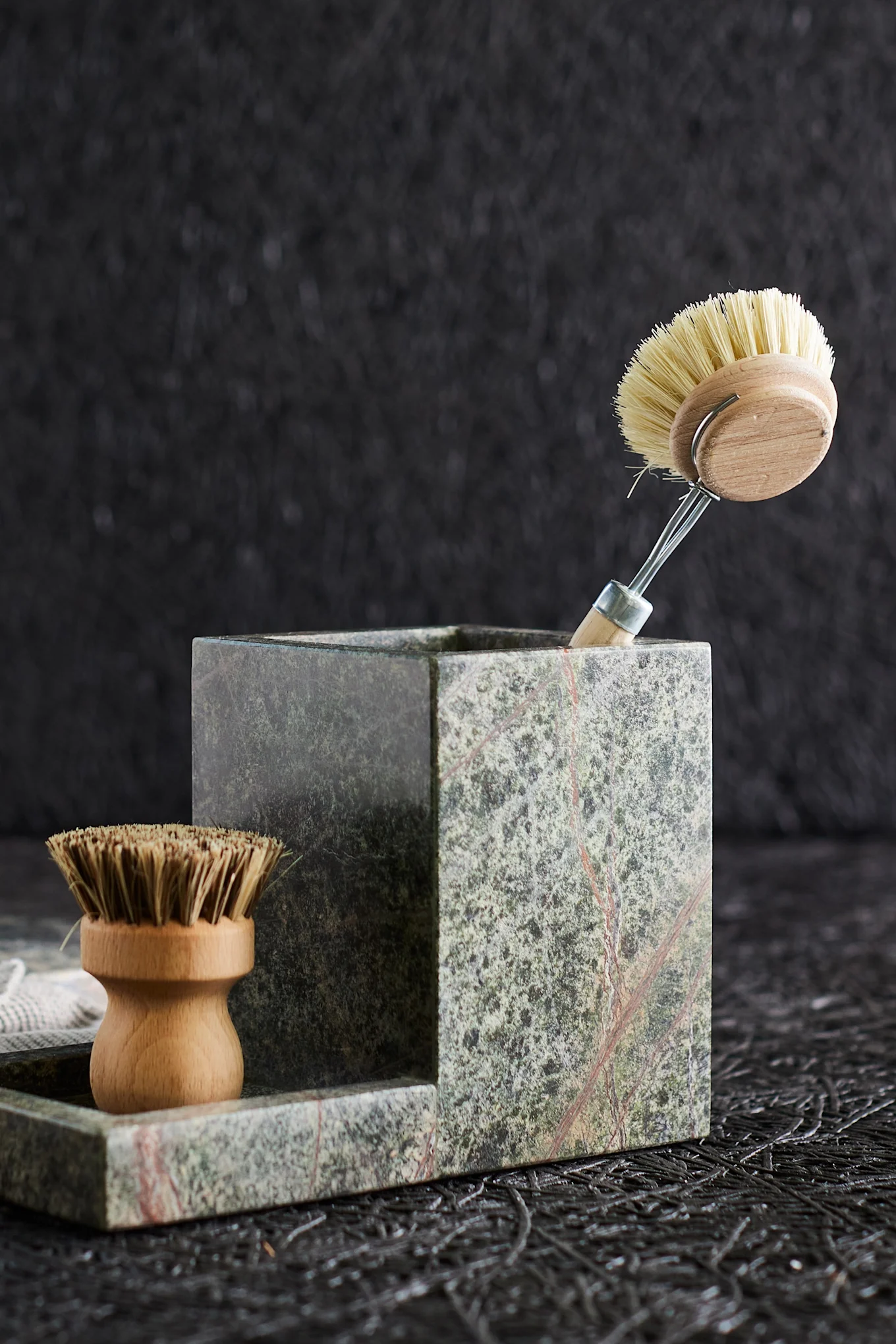 Two wooden cleaning brushes in a green marble organizer, one upright and one in a tray, on a dark background.