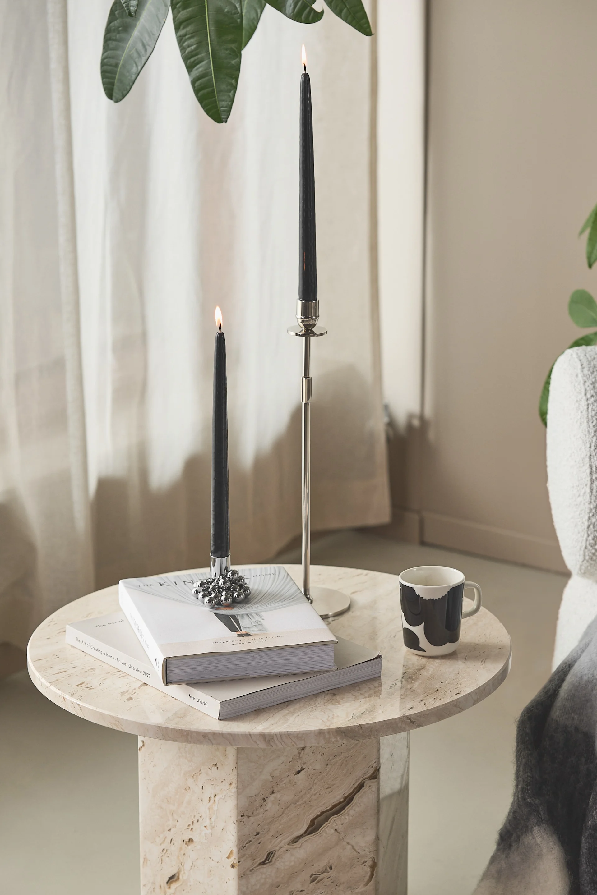 Two lit black taper candles in silver holders, a stack of books, and a patterned mug on a hexagonal travertine marble side table.