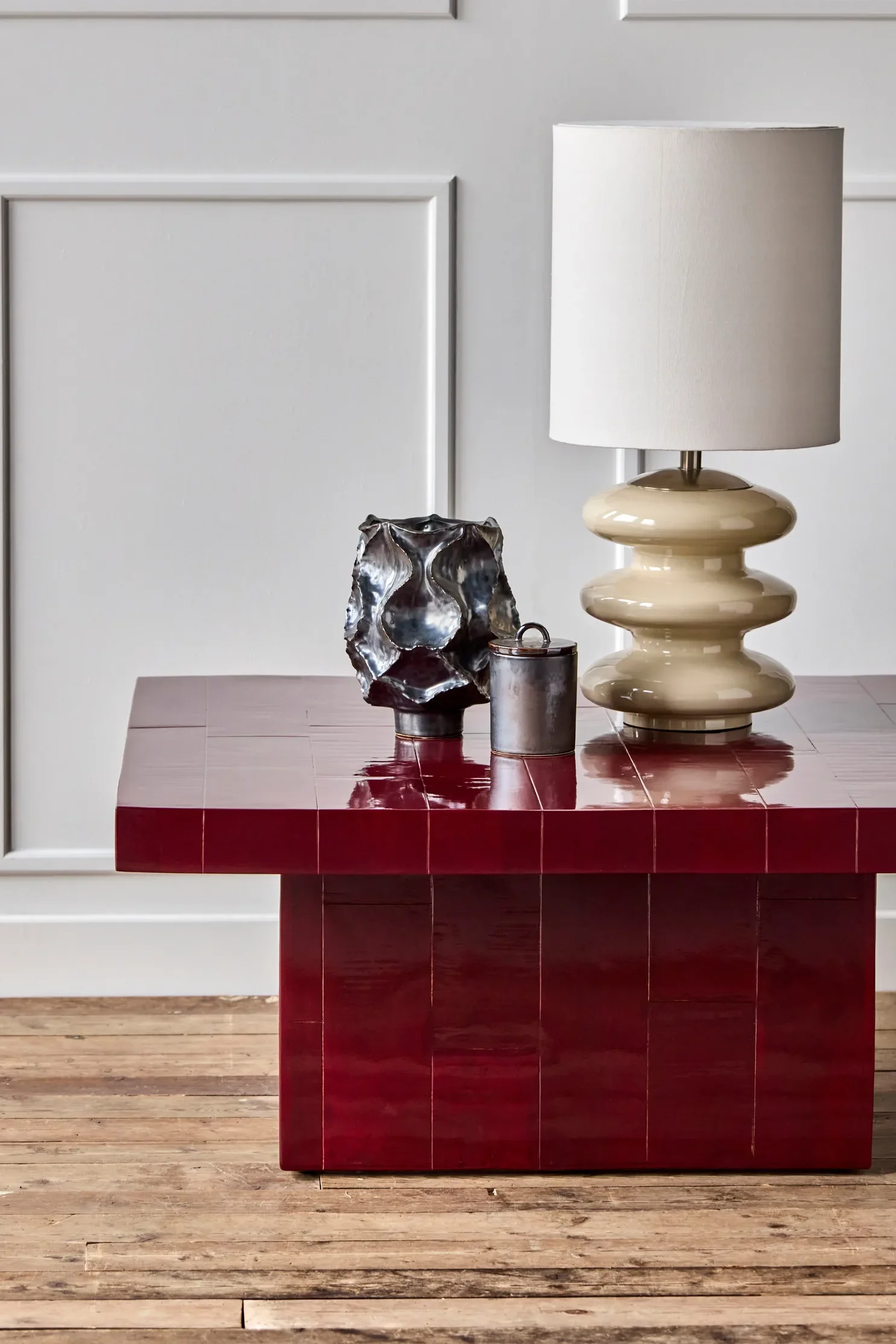 Glossy dark red tiled coffee table with a cream sculptural lamp, a dark metallic abstract vase, and a small cylindrical container.