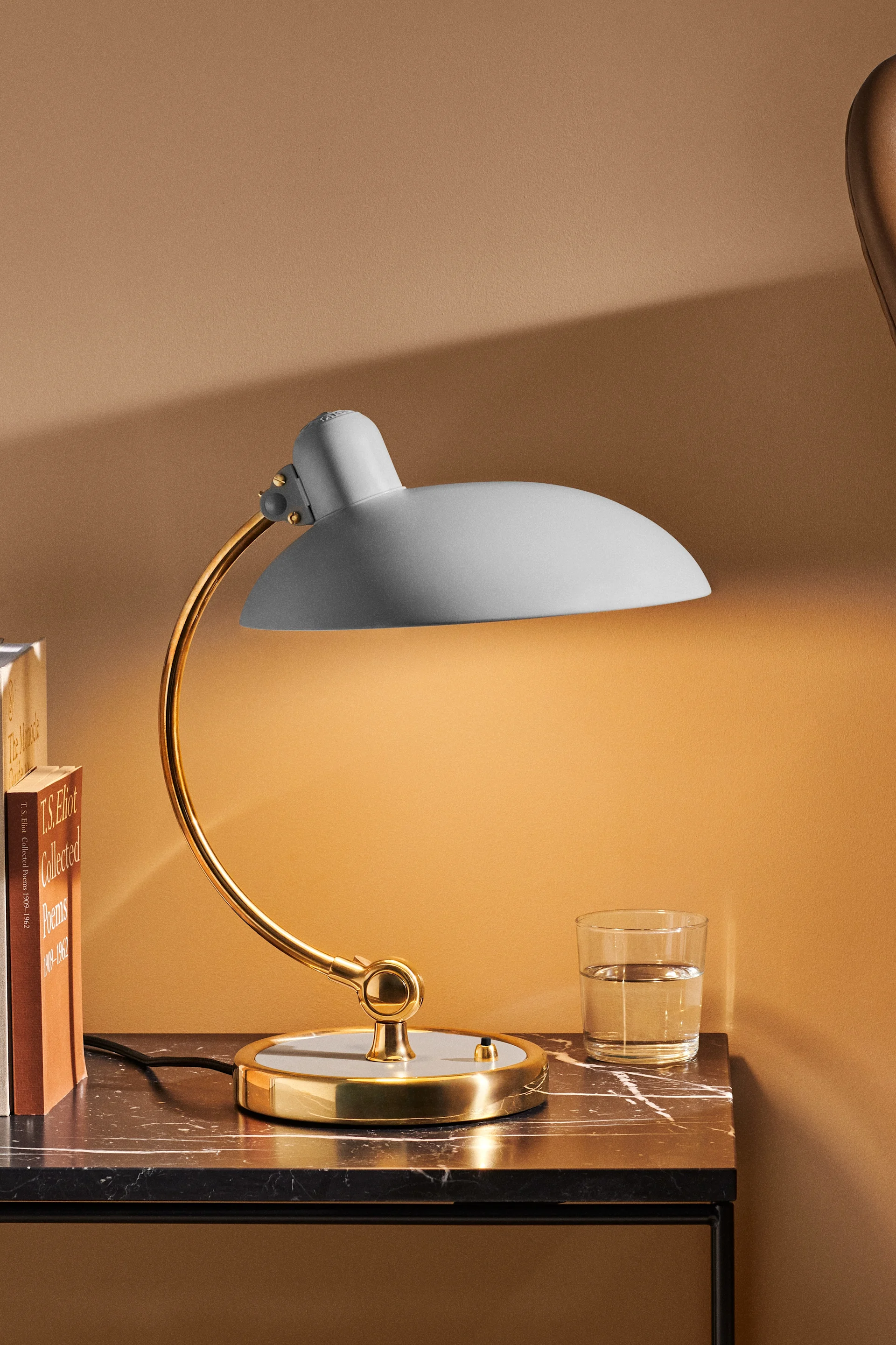 A gold and light gray desk lamp with a curved arm on a dark marble table next to books and a glass of water.