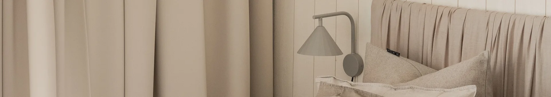 A neutral-toned bedroom features beige curtains, a grey wall lamp, and a textured beige headboard and pillow.