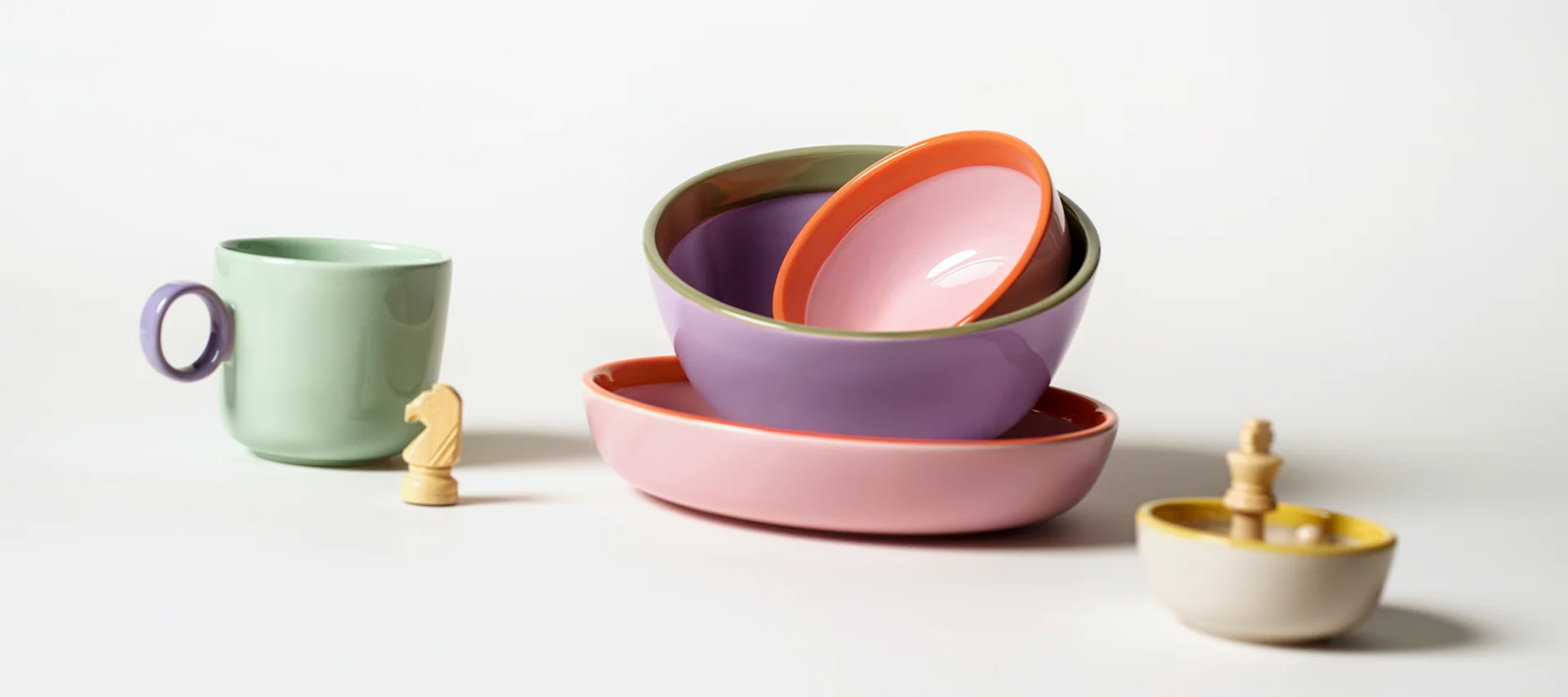 A green mug, a chess knight, and a stack of colorful ceramic bowls and plates on a white surface.