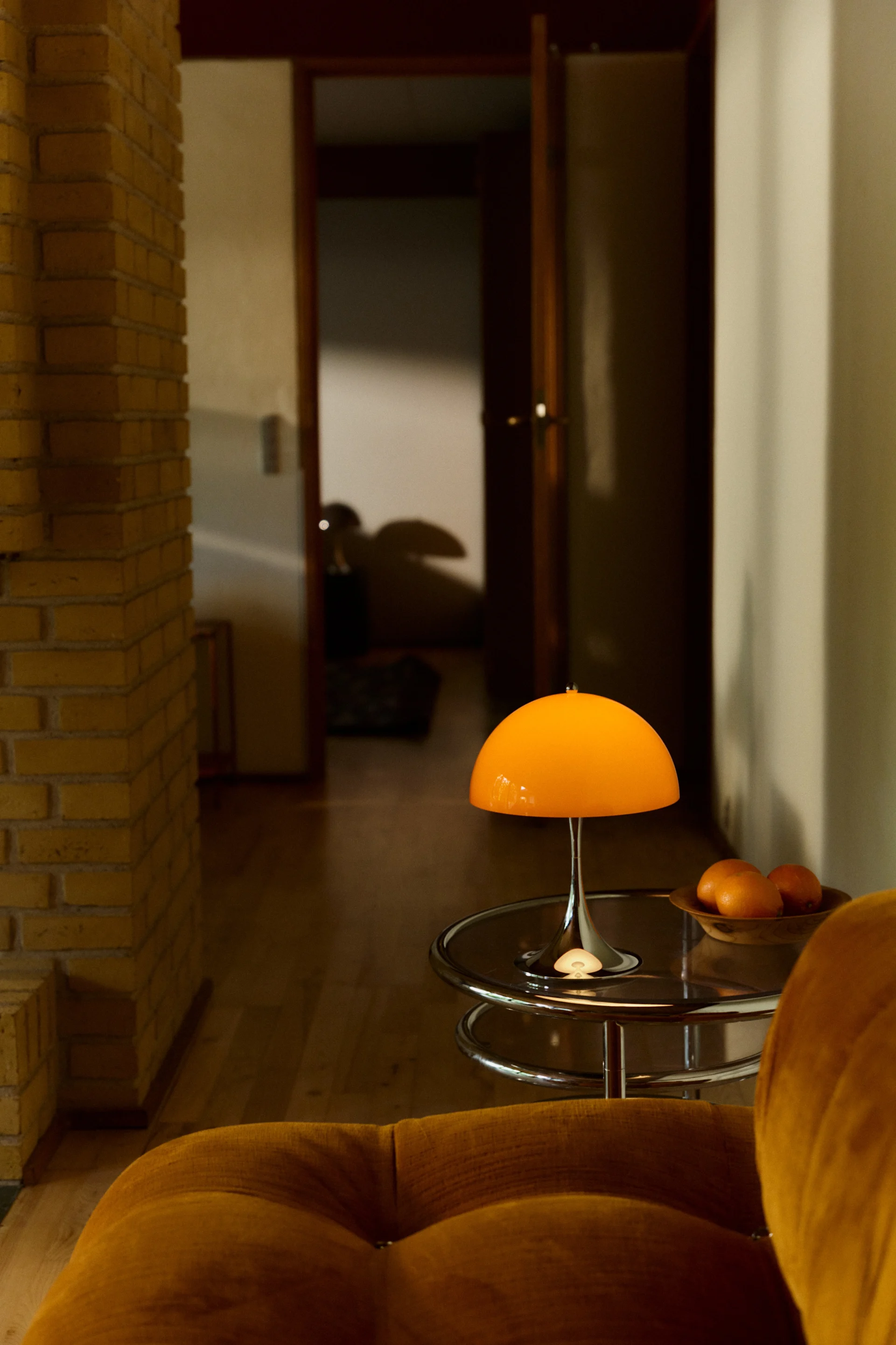 An orange mushroom-shaped lamp sits on a round glass table next to a gold velvet armchair, with a brick wall and open doorway in the background.