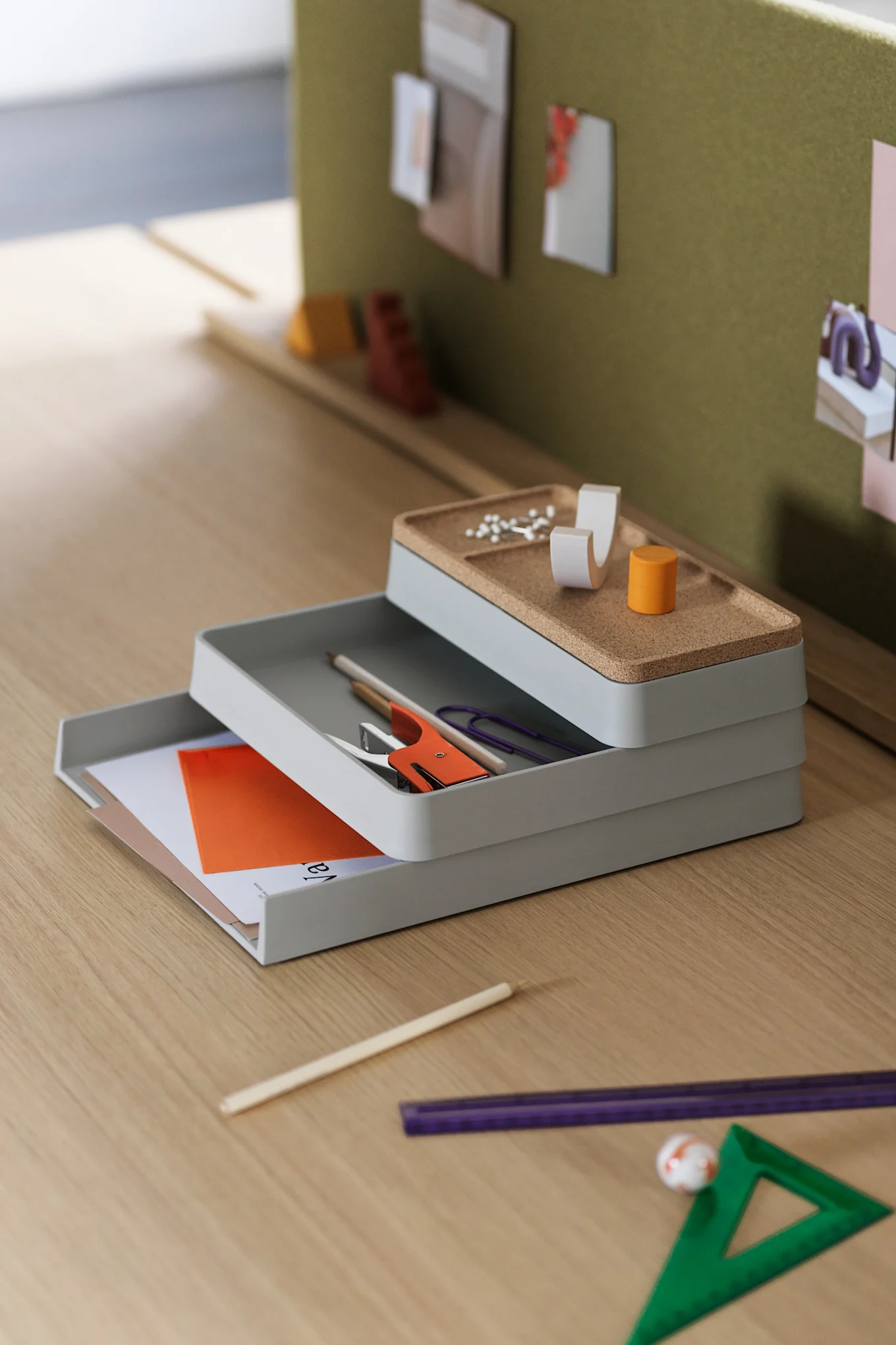 Stack of three gray desk organizers with cork-lined top tray, holding office supplies like stapler, pins, and papers on a wooden desk.