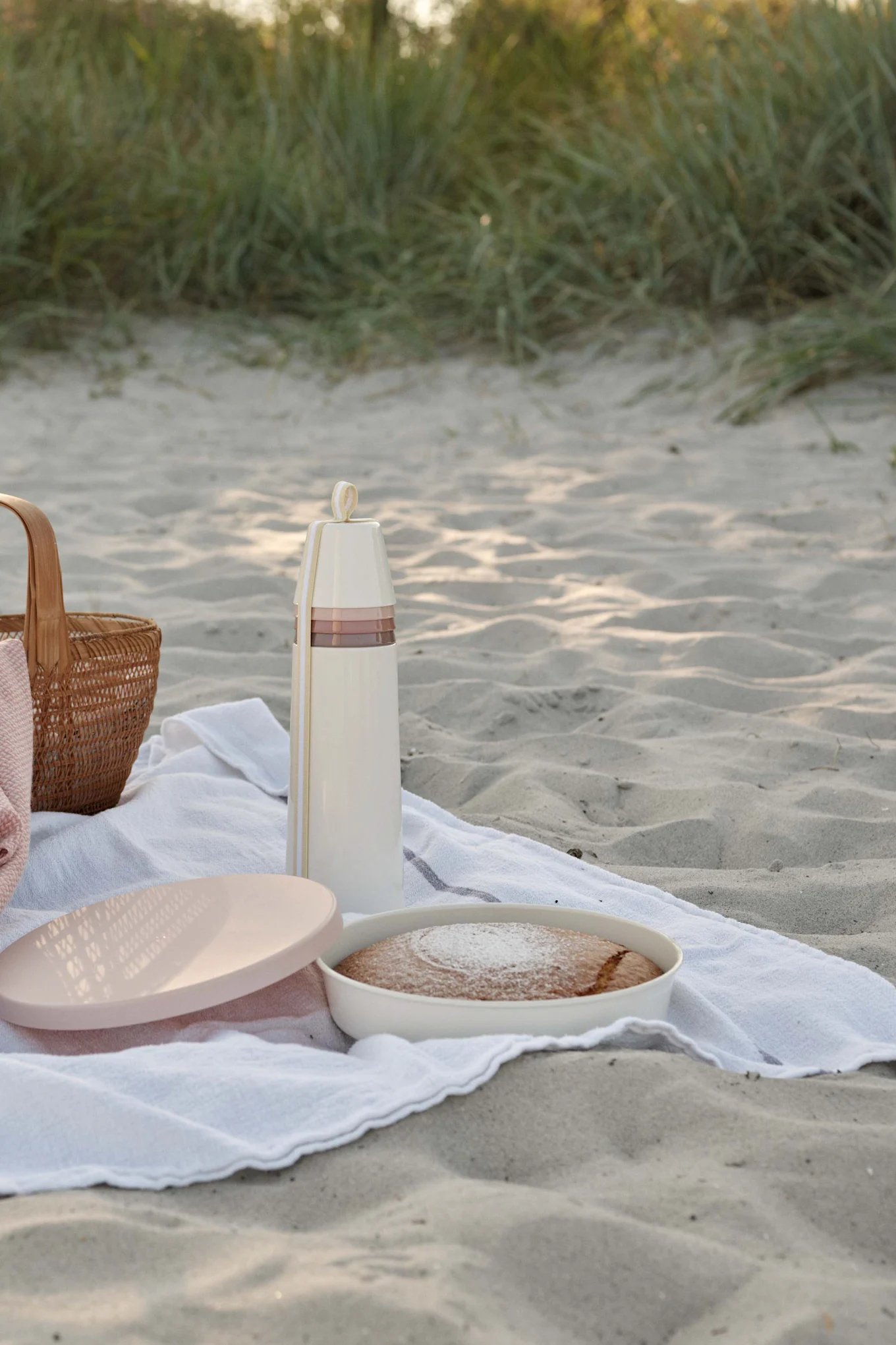 Picnic on a sandy beach with a cream thermos, cake with powdered sugar, and a woven basket.