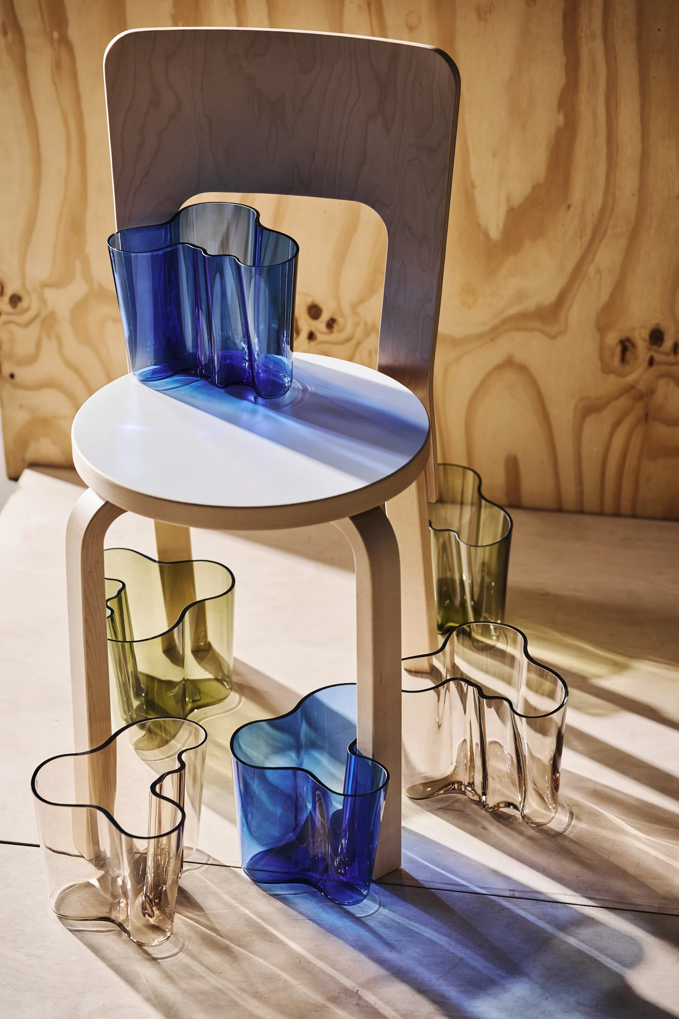 Multiple wavy glass vases in blue, olive green, and clear, arranged on and around a light wooden stool with a wooden chair in the background. Sunlight casts colored shadows.