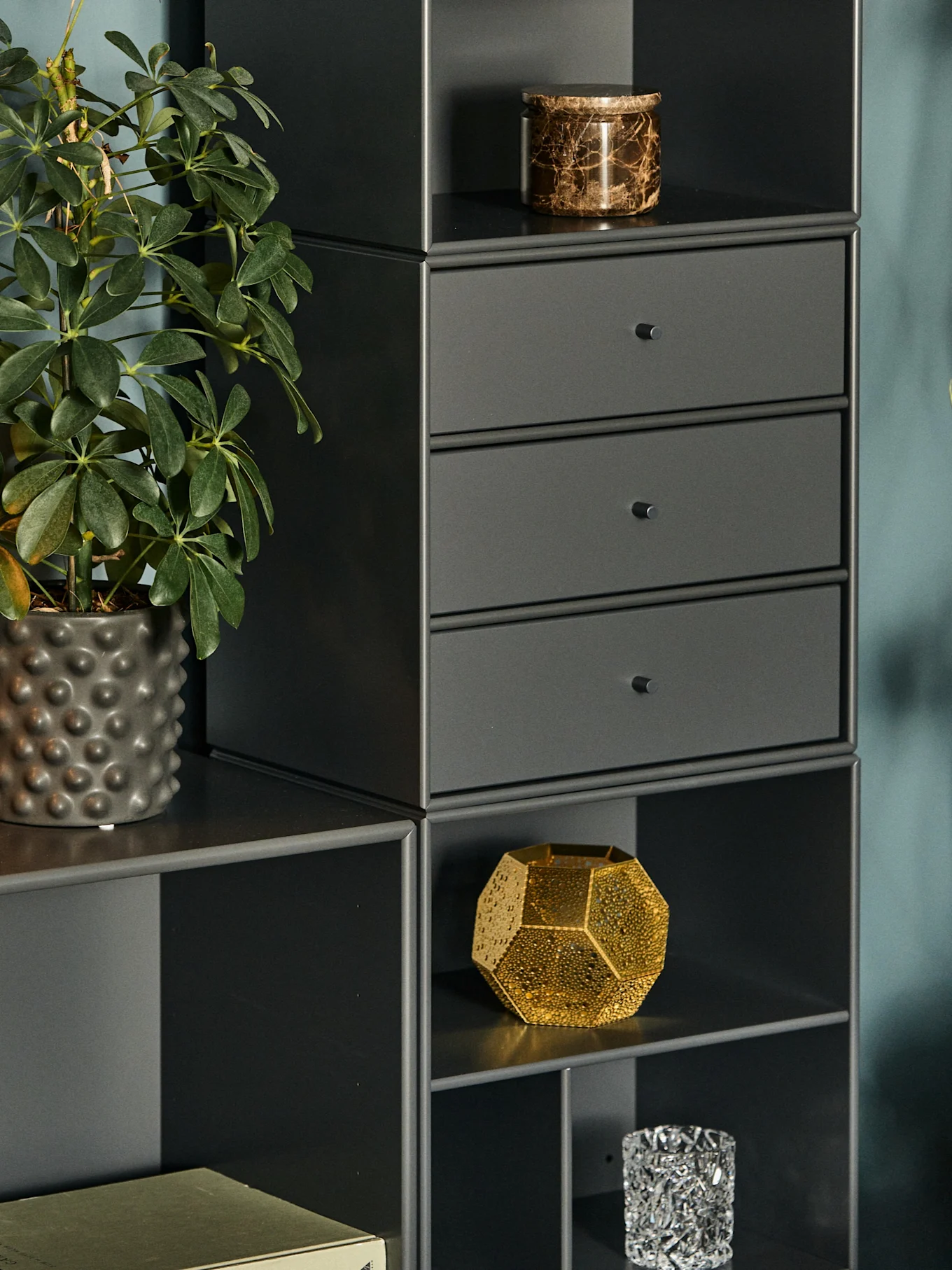 Close-up of a dark gray modular shelving unit with a potted plant, a marble jar, drawers, and decorative objects.