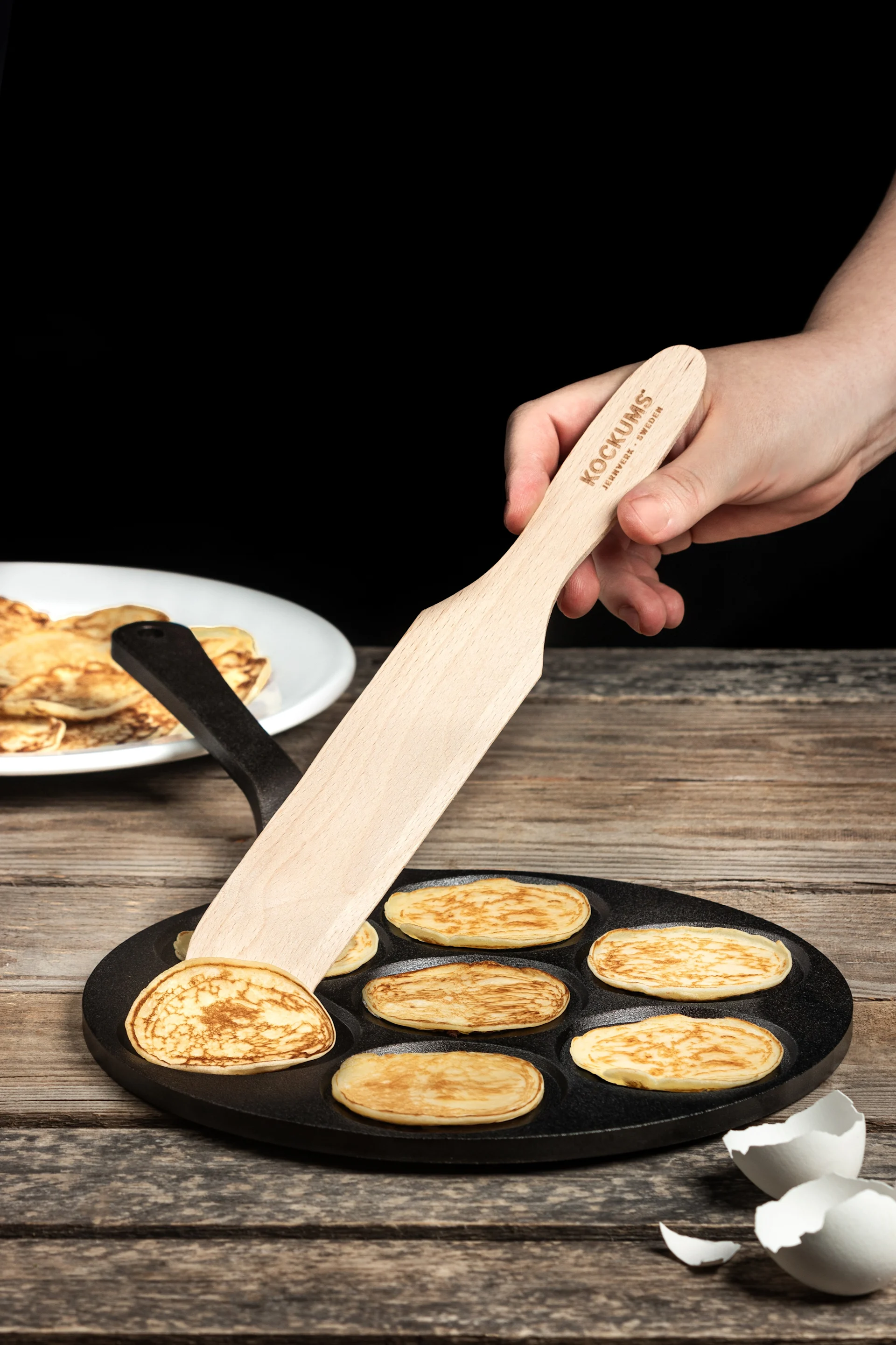 Hand using a wooden spatula to flip small, round pancakes in a cast iron pan, with more pancakes on a plate and eggshells nearby.
