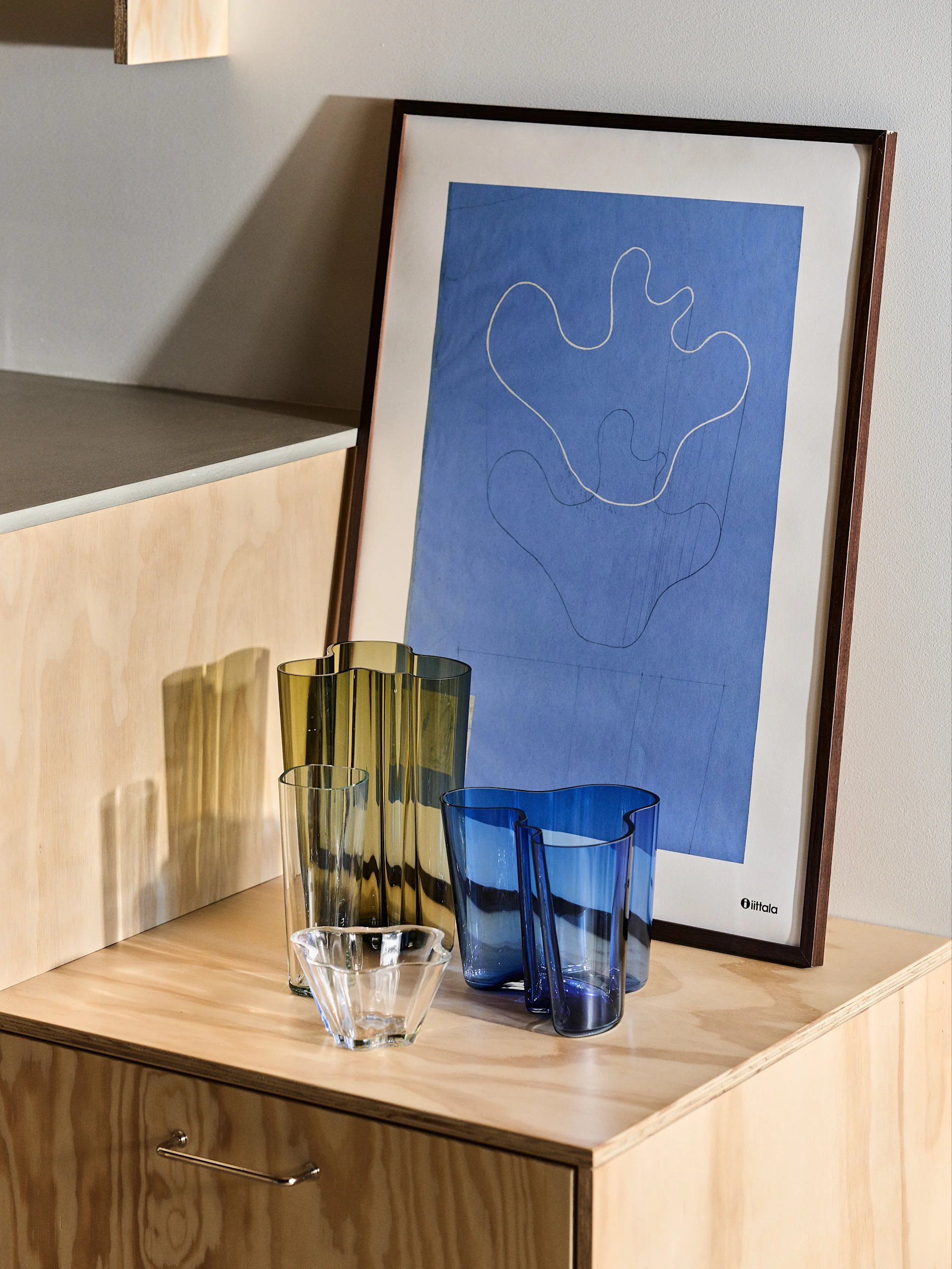 Wavy olive green and blue glass vases, clear bowl, blue tumblers on a wooden cabinet. Abstract blue Iittala framed print.