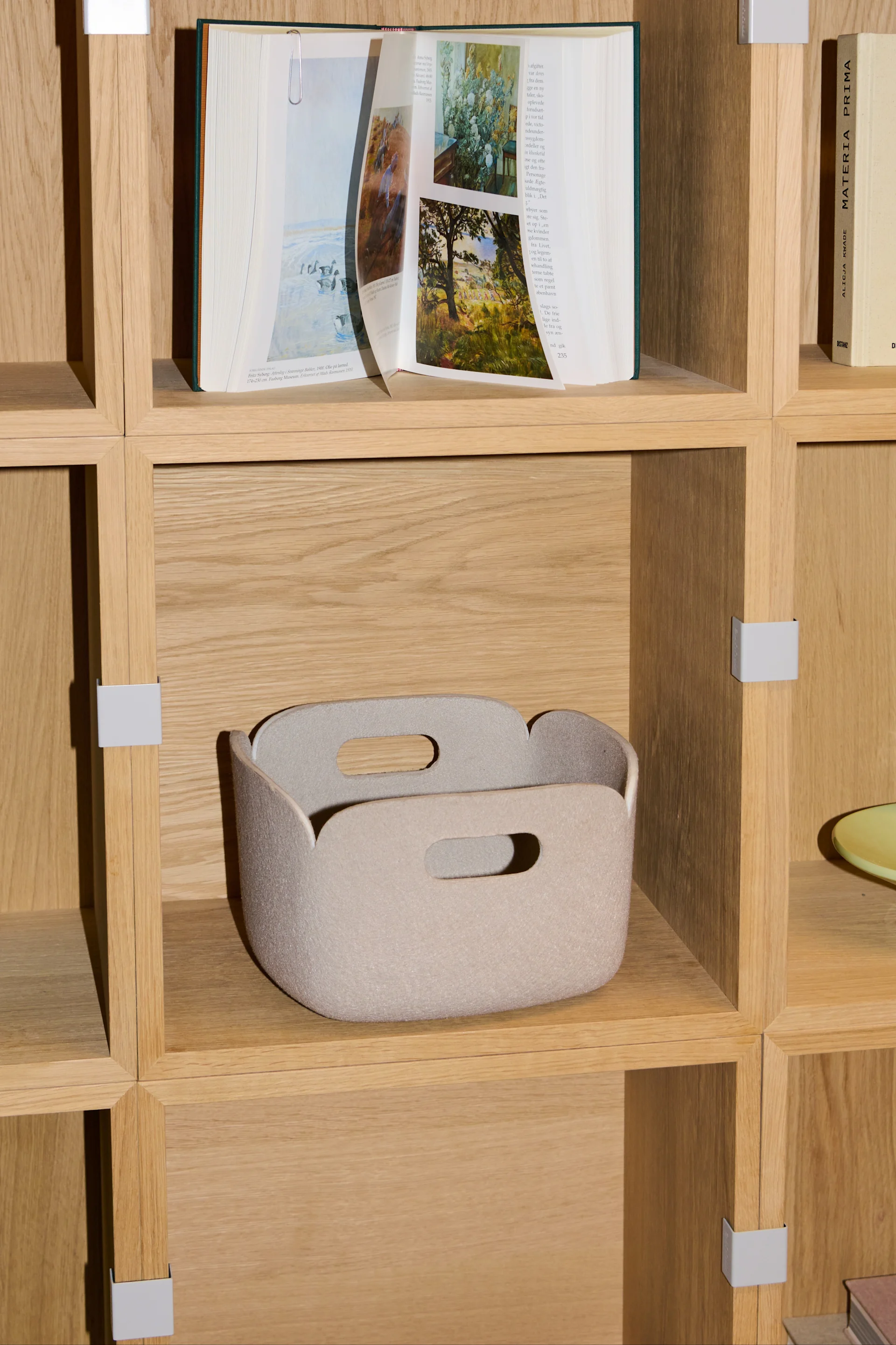 A light beige felt storage basket with two handles sits on a wooden shelf, below an open book.