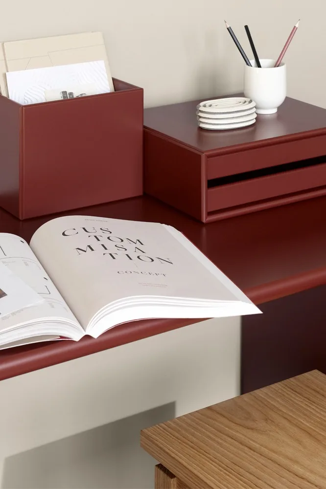 A reddish-brown desk with a file box, drawer unit, pencil holder, coasters, and an open book. A wooden stool is partially visible.