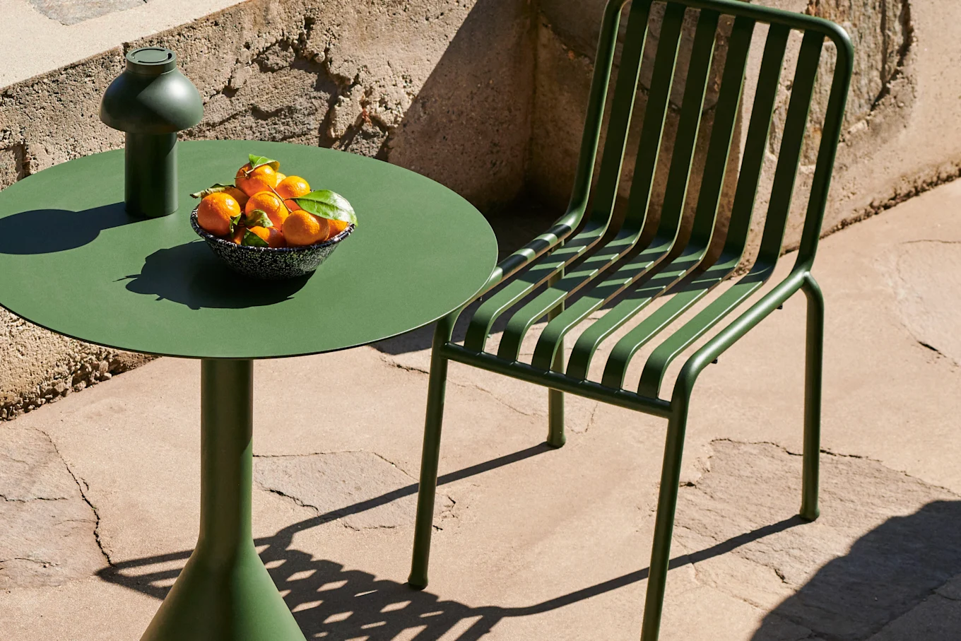 Dark green outdoor table and slatted chair on a patio, with a mushroom-shaped lamp and a bowl of tangerines on the table.