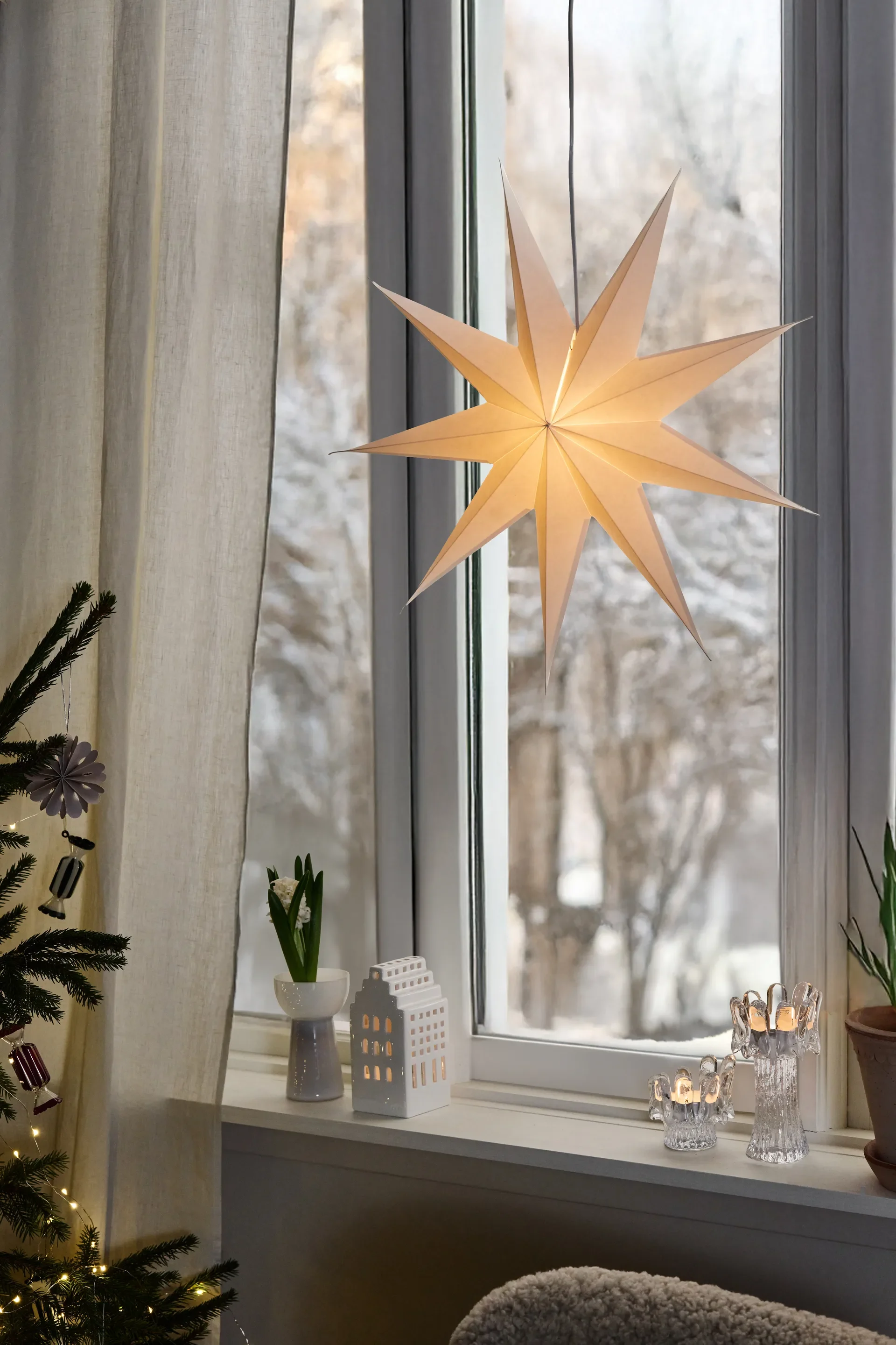 A window decorated for winter with a glowing paper star, a ceramic house, lit candle holders, and a Christmas tree.