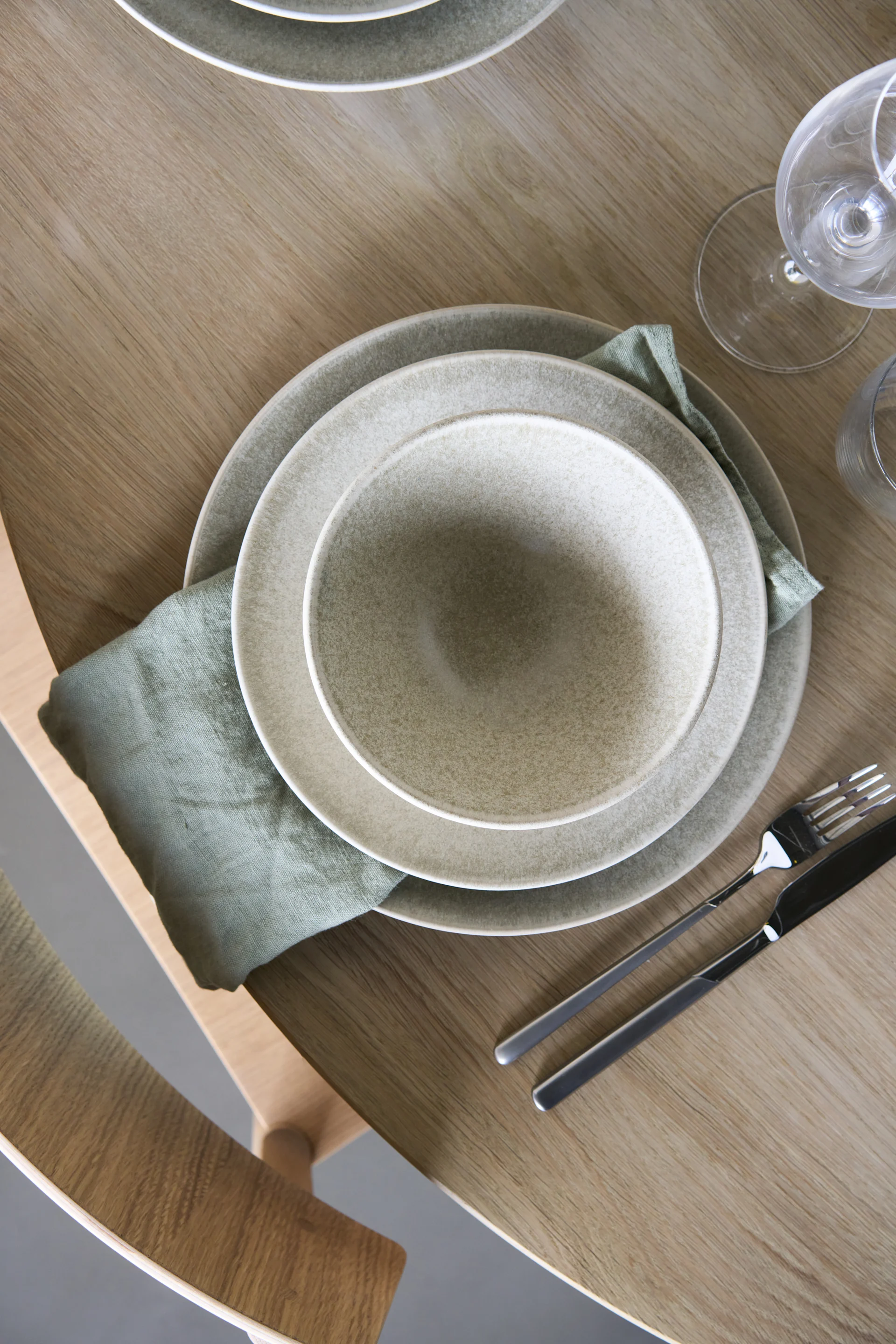 Overhead view of a dinner place setting with speckled ceramic plates, a green linen napkin, and cutlery on a light wood table.
