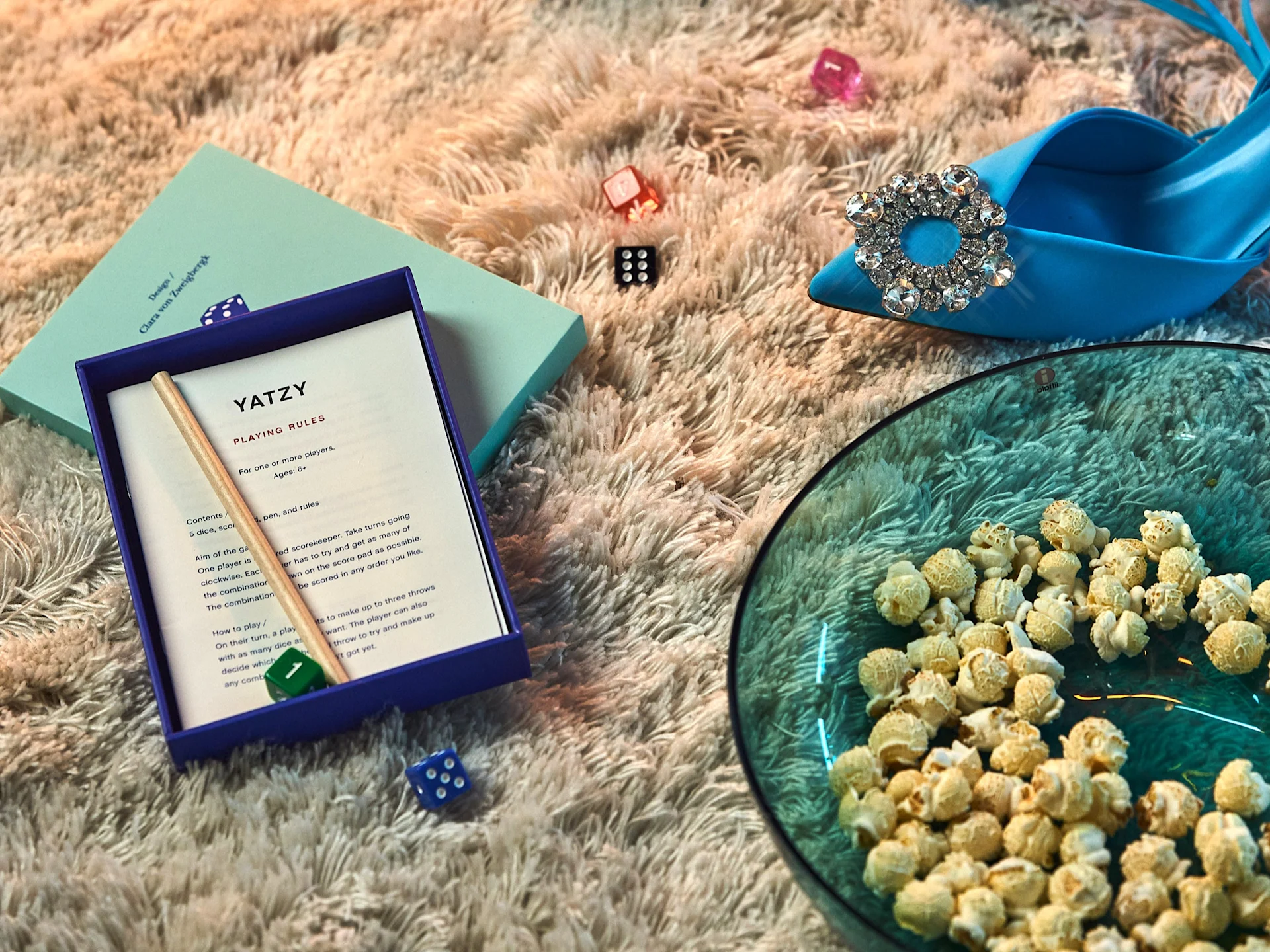 A Yatzy game box, scattered dice, a blue high-heeled shoe with a sparkly buckle, and a bowl of popcorn on a fluffy rug.