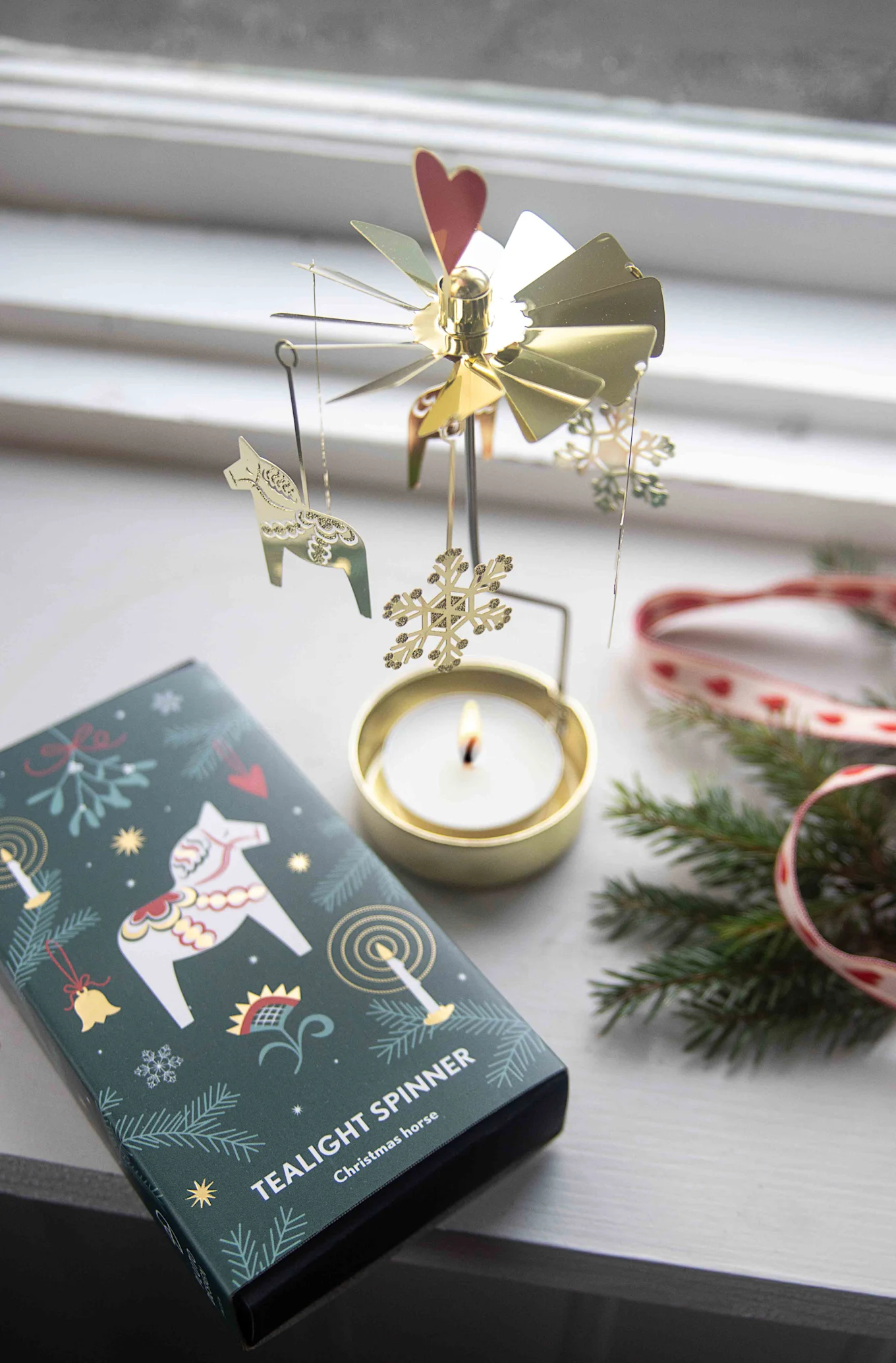 A lit golden Christmas tealight spinner with Dala horses and snowflakes, beside a matching box and fir branches.