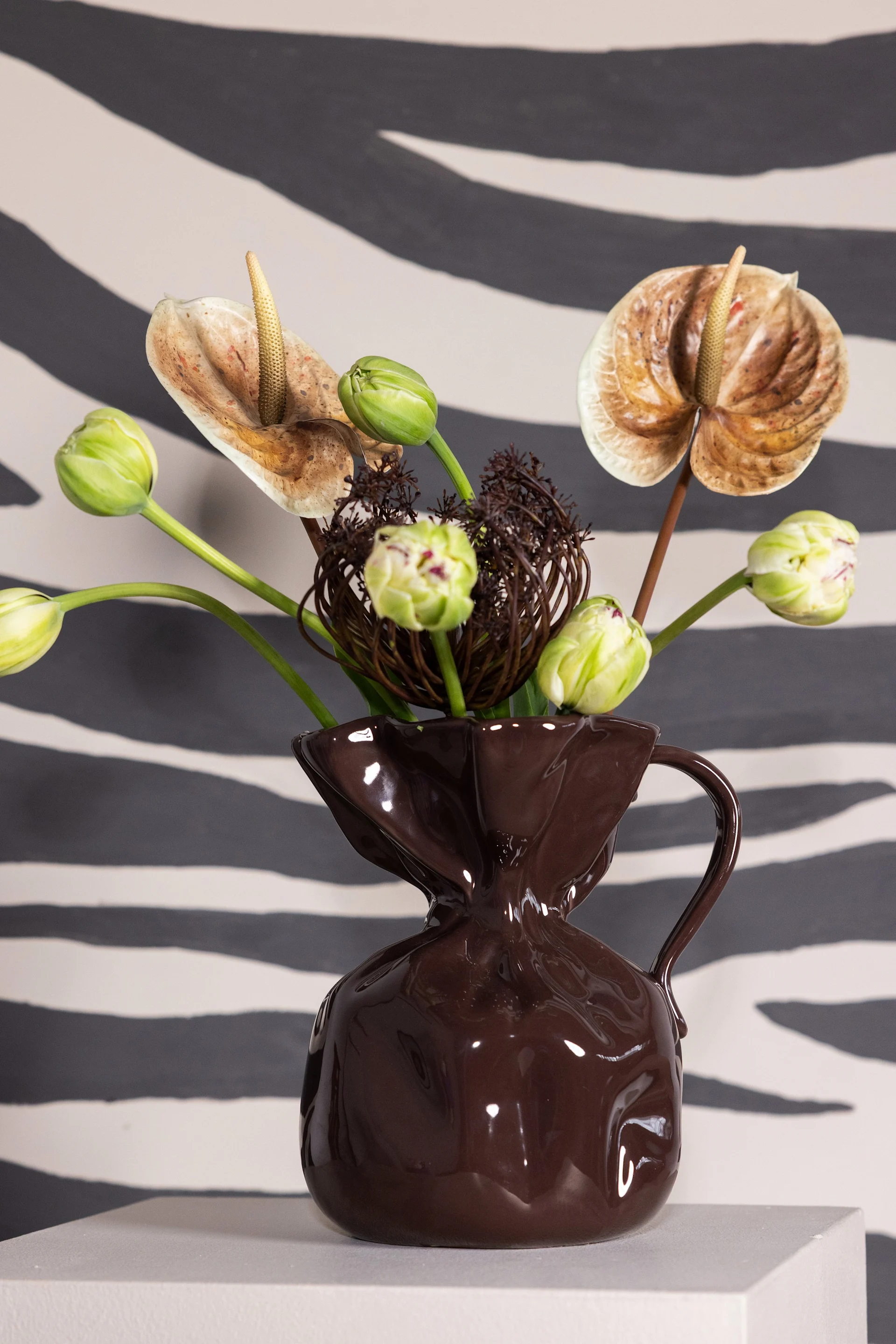 Floral arrangement in a unique dark brown vase with anthuriums, green tulip buds, and dried elements, against a zebra-stripe background.