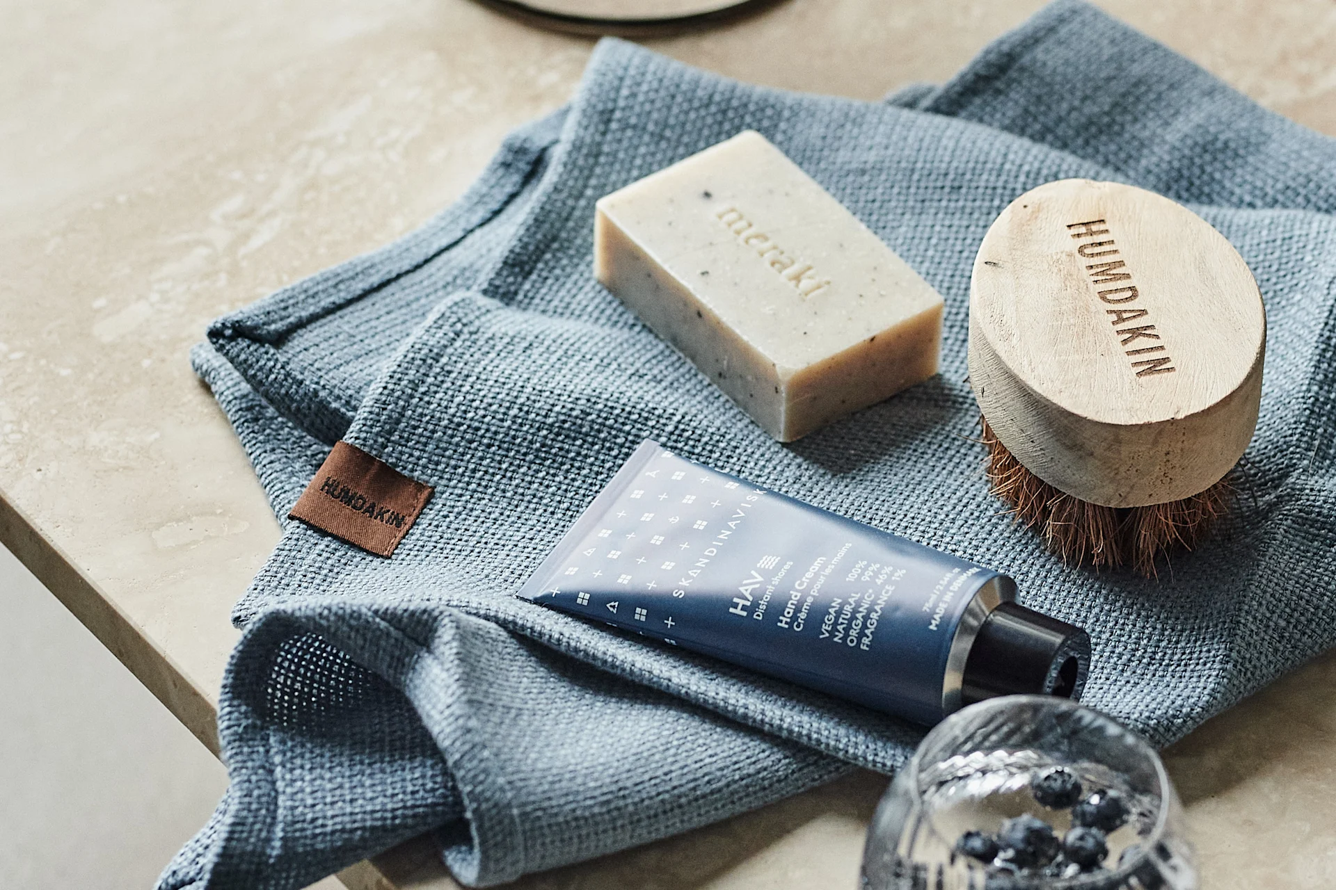 A blue textured towel, a Meraki soap bar, a Humdakin wooden brush, and Skandinavisk hand cream tube on a light countertop.