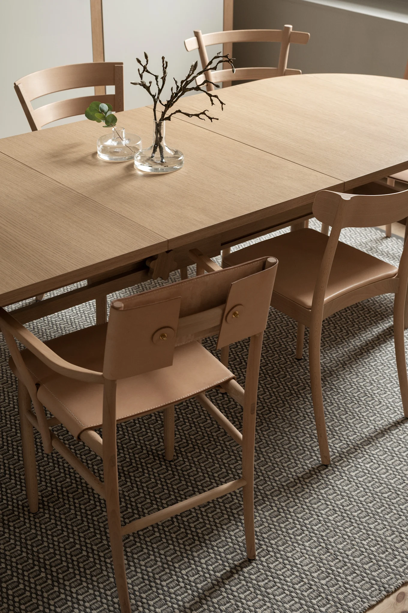 Light wooden dining table with leather and wood chairs on a patterned rug, decorated with plant branches in glass vases.